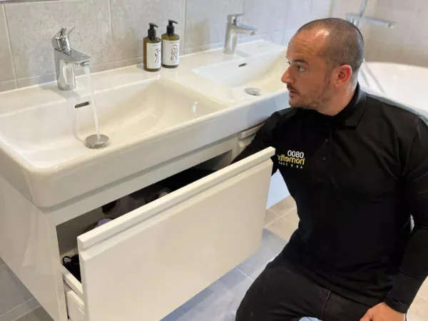 A gas safe plumbing technician inspecting under a double bathroom sink cabinet for water leaks, wearing a branded 0800 HomeFix uniform, in a modern bathroom setting